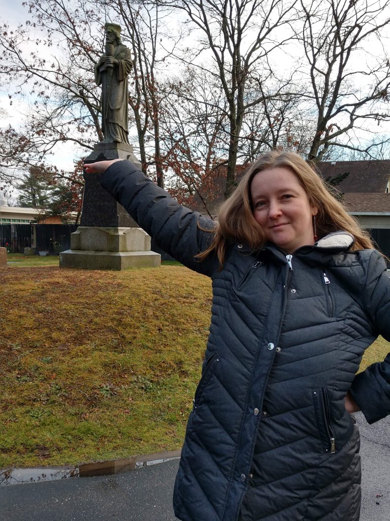 Photograph of woman and Index research staff member, Jessica Savage, with raised arm before stone statue of the medieval Bohemian martyr Jan Hus in Union Cemetery in Bohemia, Long Island, New York.
