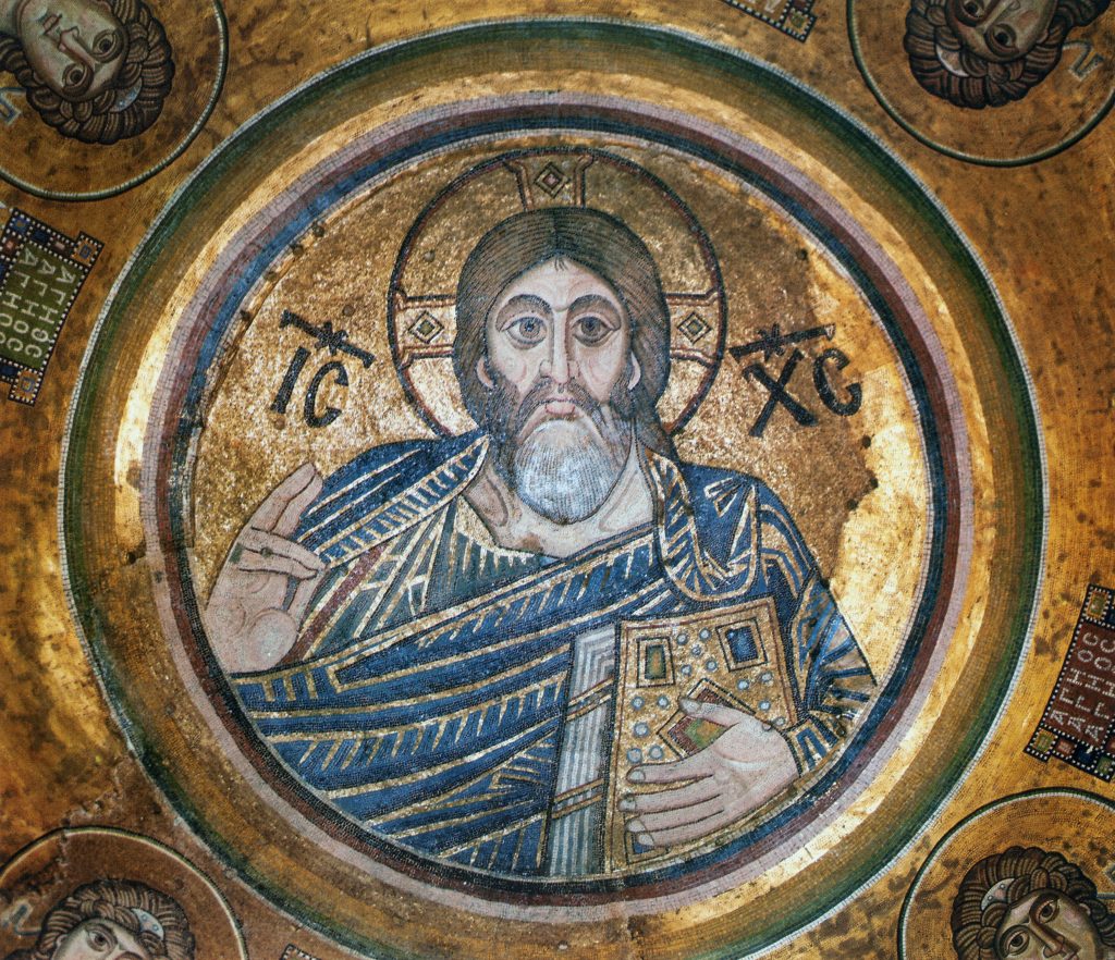 Mosaic depicting bust of bearded man wearing halo, raising right hand and holding book in left hand, against gold background and within multi-colored circular frame.