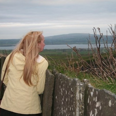 A woman with long blond hair, wearing a yellow jacket, looking over a stone wall into a green landscape with hills beyond water in the background.