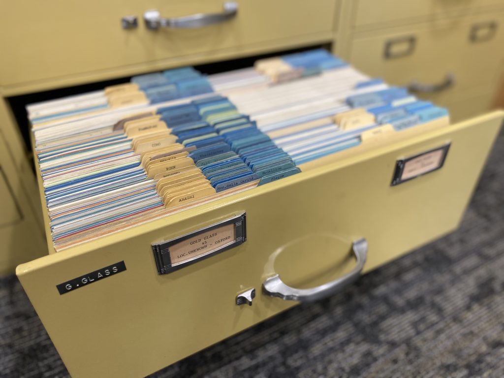One open yellow file cabinet drawer, labeled “G. Glass,” containing yellow and blue-tabbed dividers and two rows of cards.