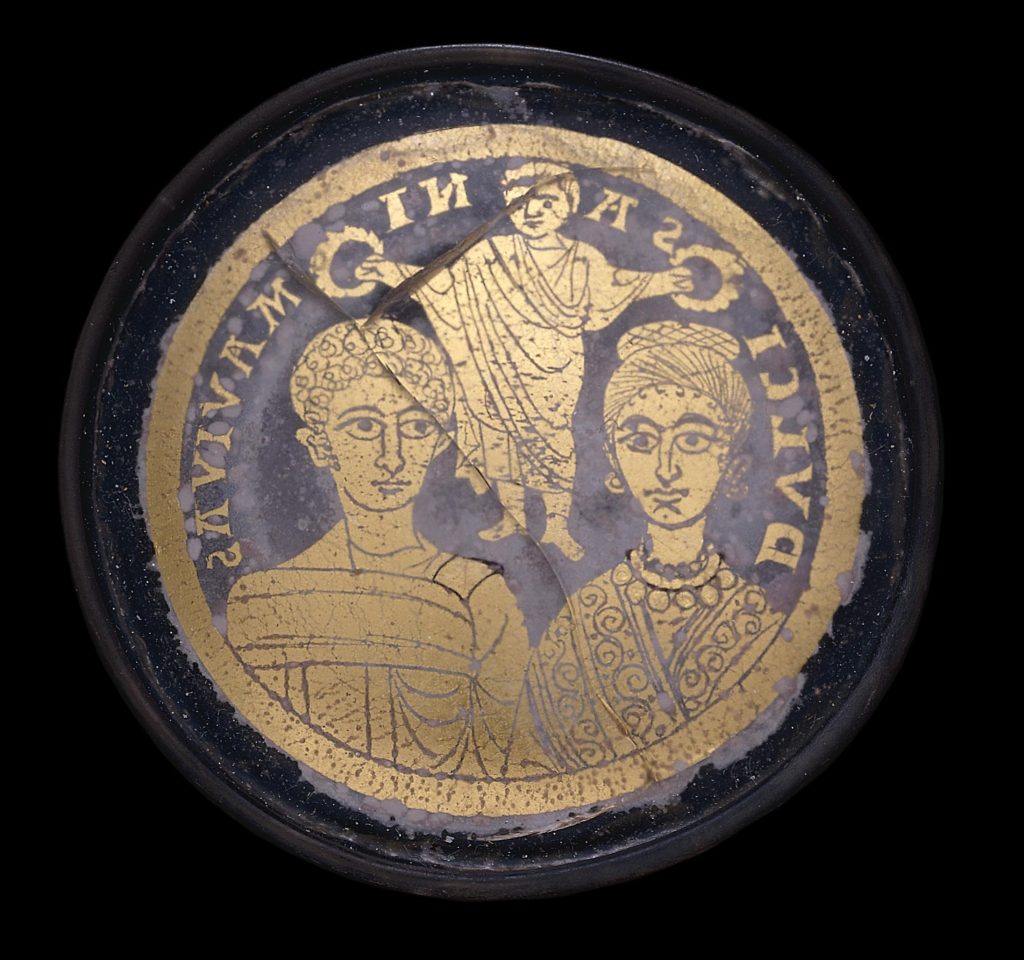 A glass medallion decorated in gold with the bust portraits of a man and woman next to one another, both of their heads crowned with wreaths held in the hand of a man partly draped in a garment between them. Latin inscription around the upper edge of the medallion, in reverse: DVLCIS ANIMA VIVAS.