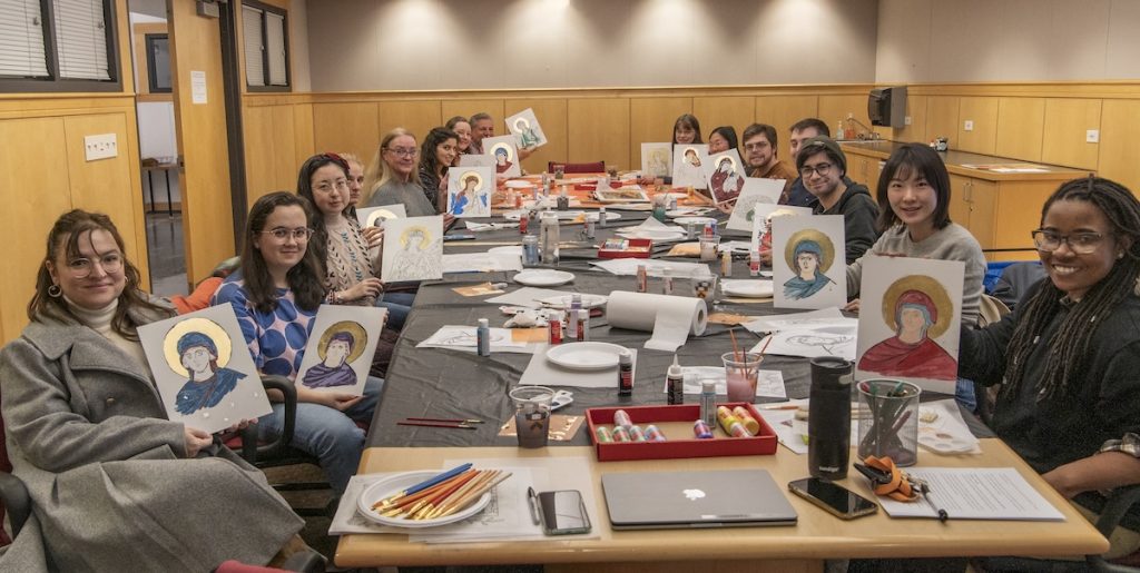 About fifteen students surround a long table covered in art supplies, and each student holds up a small panel containing their artwork and icon painting.