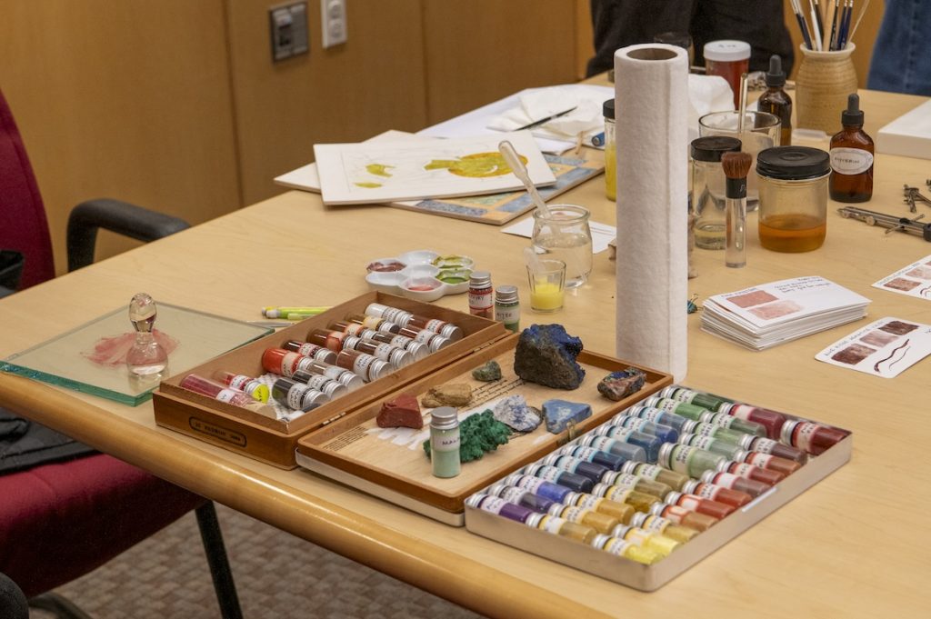 A table spread with various art supplies, paper towel roll, brushes, and sheets.