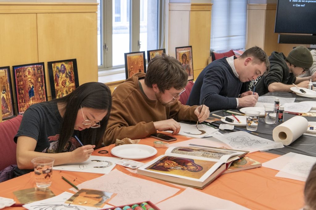 Four participants seated at a table spread with art supplies, bend over their panels with brushes in hand. More icon reproductions leaning against the wall in the background.
