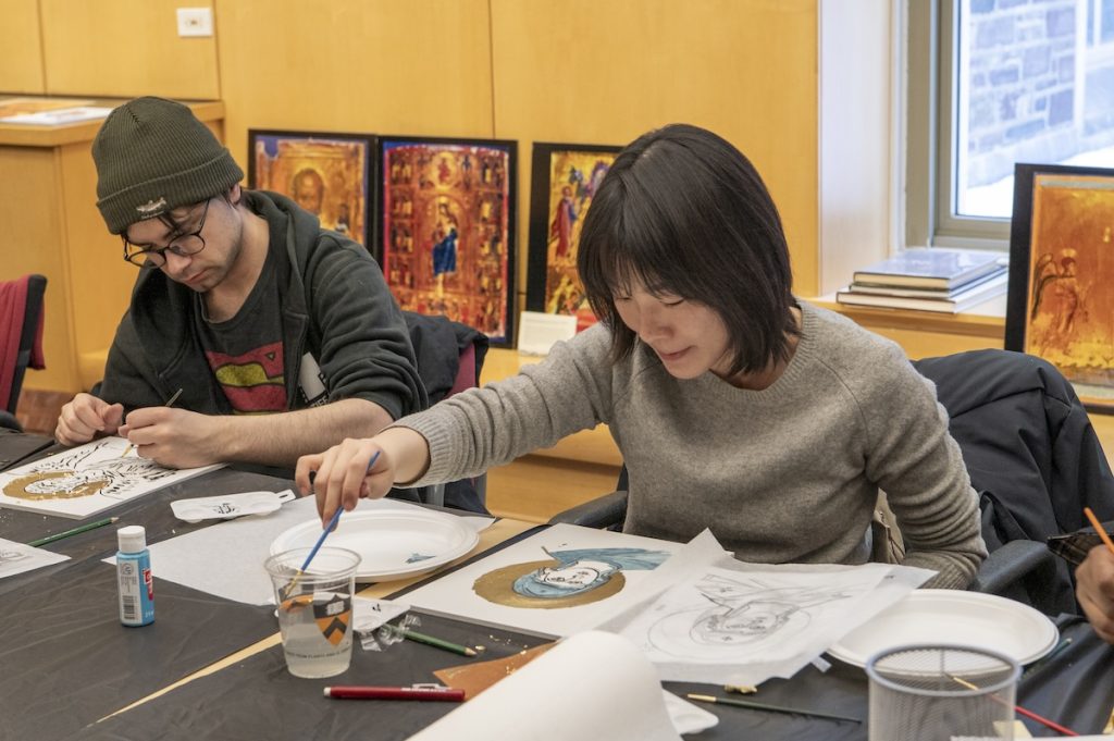 Two participants, holding art supplies, lean over their panels while making works of art. More icon reproductions leaning against the wall in the background.