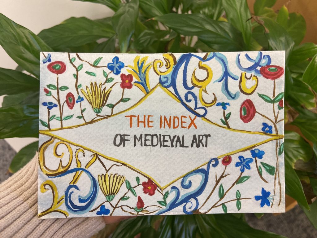 A rectangular card painted with the text “The Index of Medieval Art” within an elongated diamond frame and surrounded by red, yellow, and blue floral ornament and scrolls. Plant leaves in the background.