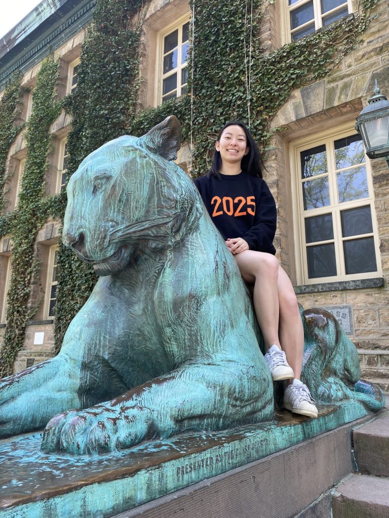 A woman, wearing a dark shirt with the orange number 2025, seated sideways on an oxidized bronze statue of a tiger in front of a brick building with windows and ivy growing up the walls.