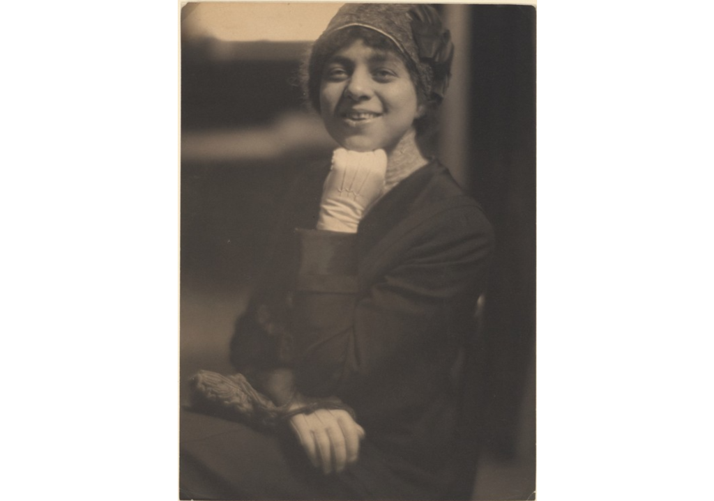 A portrait of a young woman smiling while resting her chin on her left hand. She wears a cloche hat and white gloves and holds a small handbag.