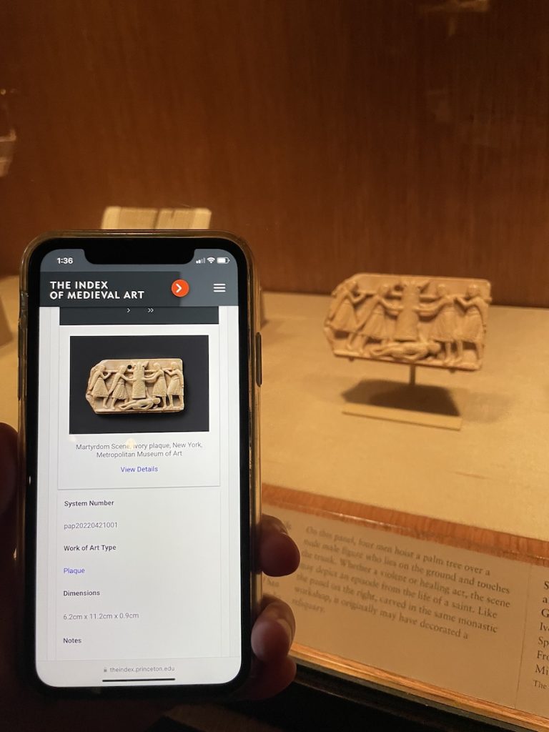 A hand holding a mobile phone showing the database record of the Index of Medieval Art as the image of an ivory object and metadata. To the right, behind museum display glass and printed label, the same ivory object is displayed on a mount.